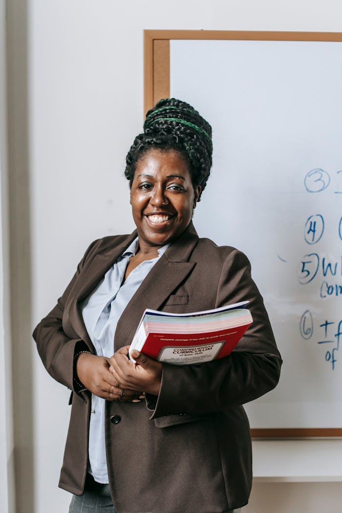 Smiling female lecturer holding books in front of classroom whiteboard.