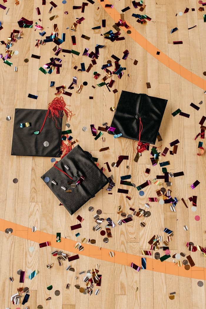 Three black graduation caps surrounded by colorful confetti on a wooden floor.